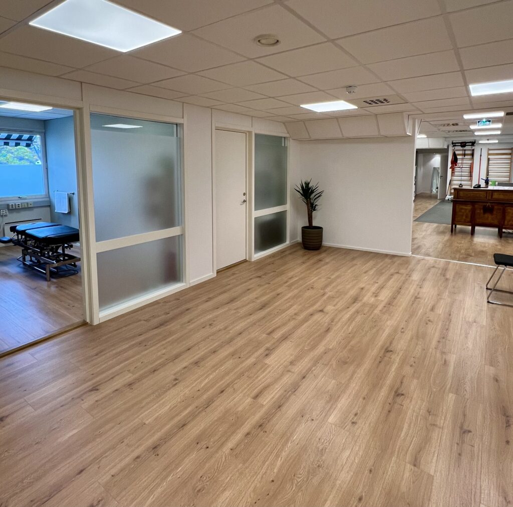 Fellesareal i klinikken med tregulv, glassvegger inn til behandlingsrom og resepsjon i bakgrunnen